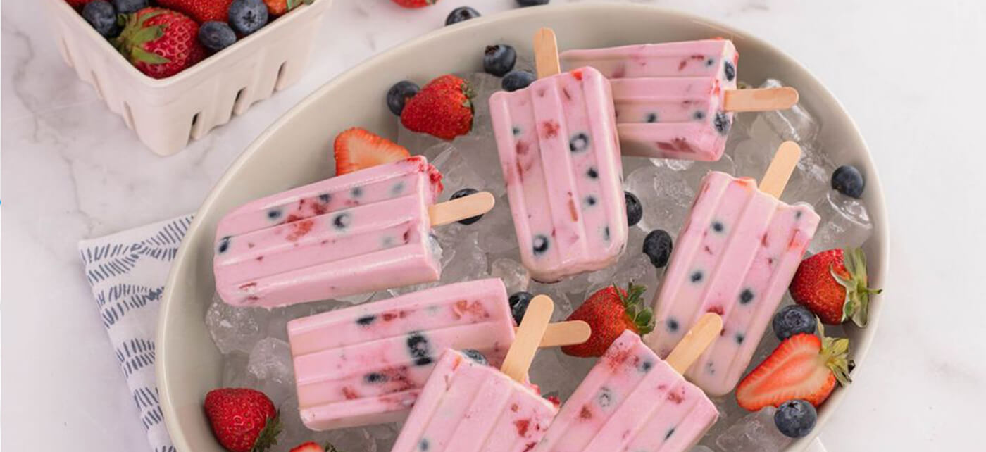 Pink creamy popsicles with mixed berries embedded inside, served on ice with fresh strawberries and blueberries