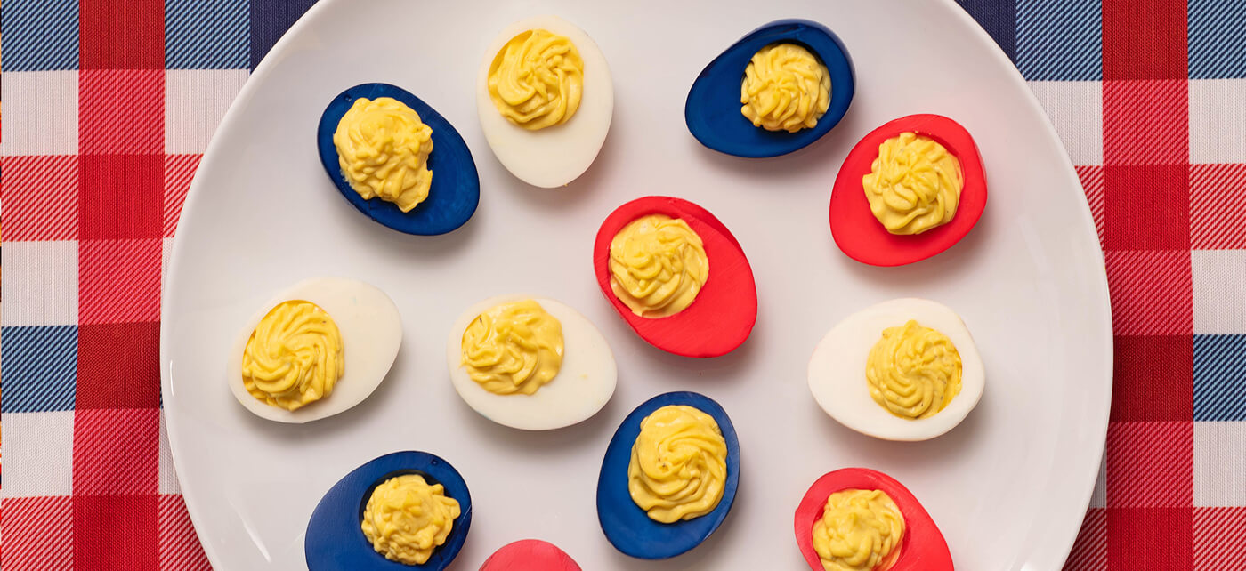 Deviled eggs dyed red, white, and blue arranged on a plate over a checkered tablecloth, topped with piped yellow filling