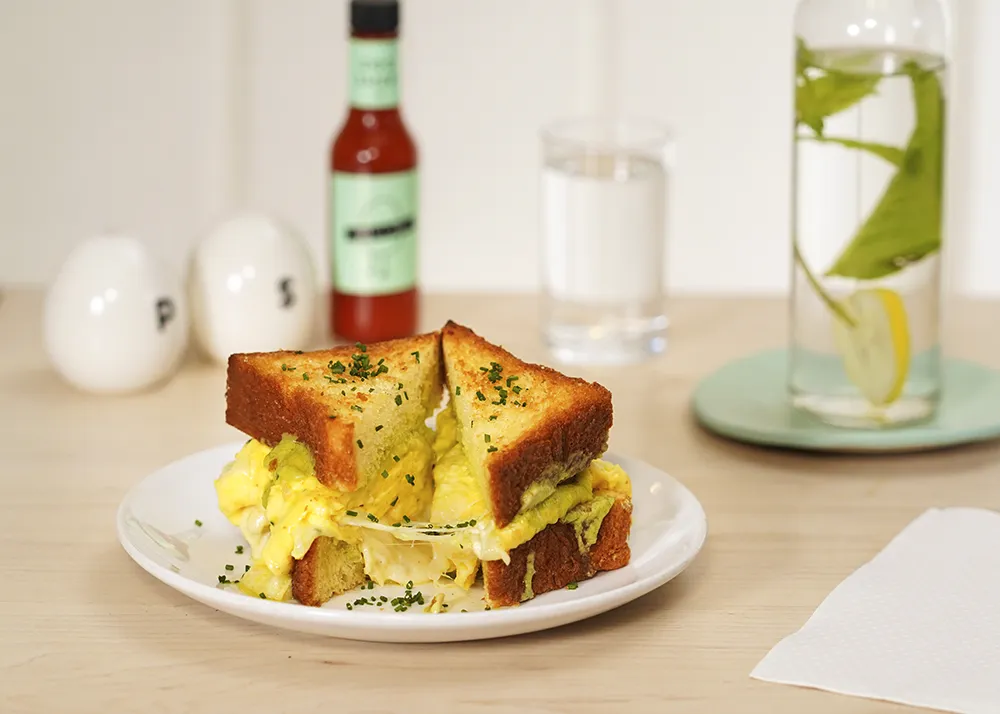 A breakfast sandwich cut in half on a white plate, filled with scrambled eggs, melted cheese, and herbs on toasted brioche bread.